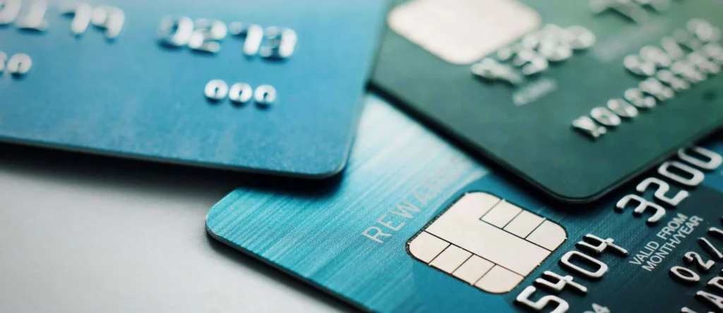 Header image depicting financial hardship, featuring a cracked credit card, stacks of overdue bills, and a calculator on a muted blue and gray background. Represents themes of debt and rising credit card defaults.